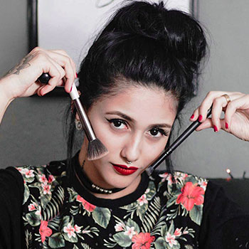 The studio owner smiling and holding two makeup brushes near her face, with bold eyeliner and red lipstick.