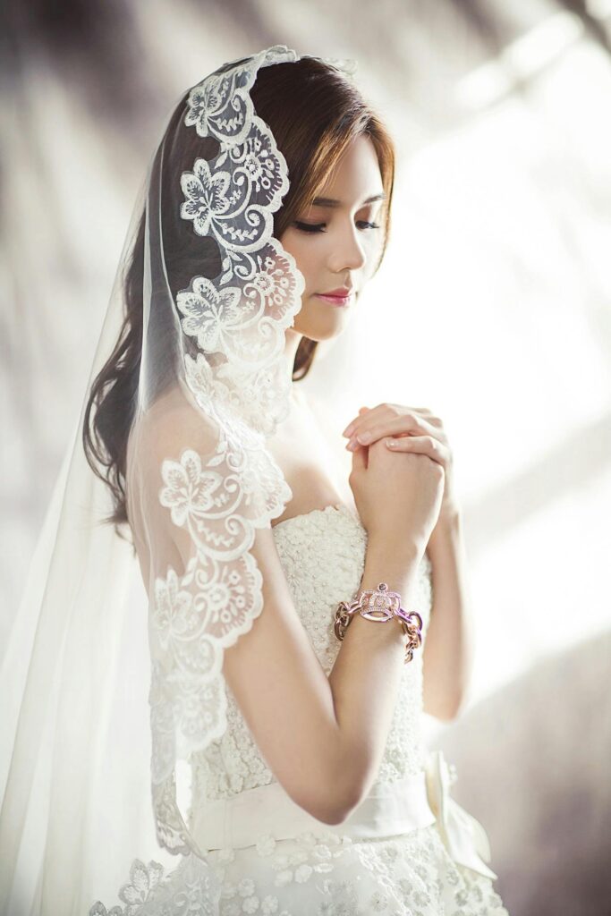 A bride wearing a white lace veil and wedding gown, posing gracefully with soft natural makeup in a bright and dreamy photoshoot setting.