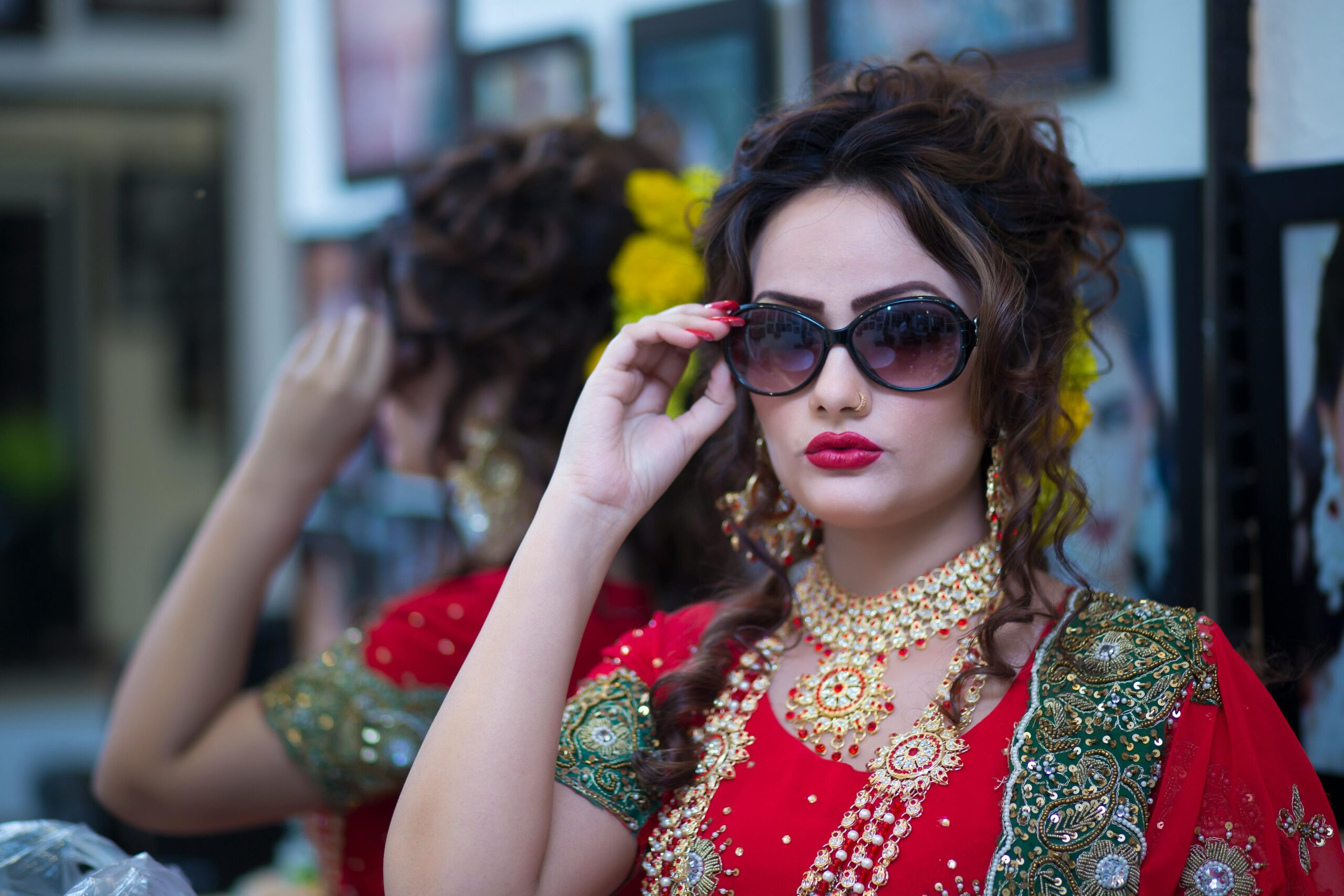 A bride wearing a red and green embroidered bridal outfit with heavy jewelry and bold makeup, posing confidently while adjusting her sunglasses in Mancherial.