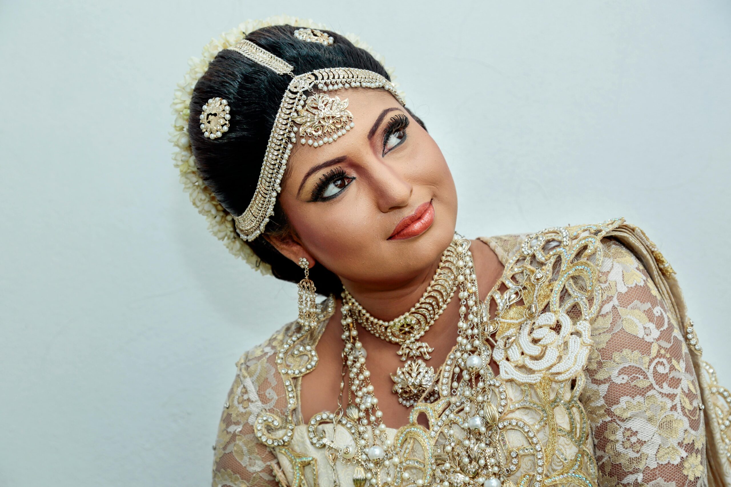 A bride wearing an ornate traditional outfit with pearl and gold jewelry, intricate lace detailing, and soft glam makeup while looking slightly upward.